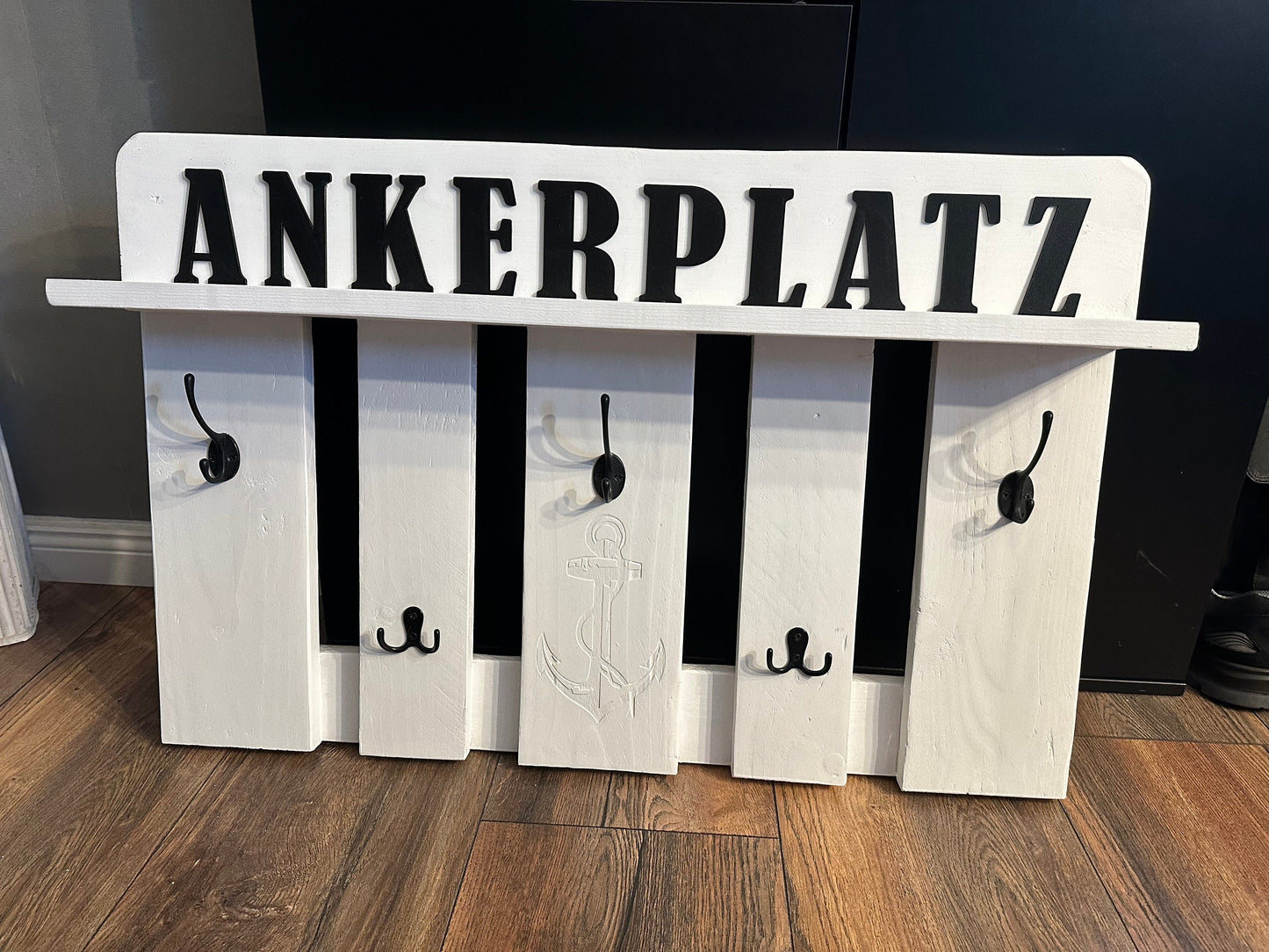 Wall-mounted wooden coat rack with five vertical slats and multiple black
metal hooks. The top board features the word “ANKERPLATZ” (“anchor place” in
German) in bold black letters, with a central anchor engraving reinforcing the
nautical theme. The rack rests on a wooden floor, evoking rustic coastal
charm and practical entryway organization.