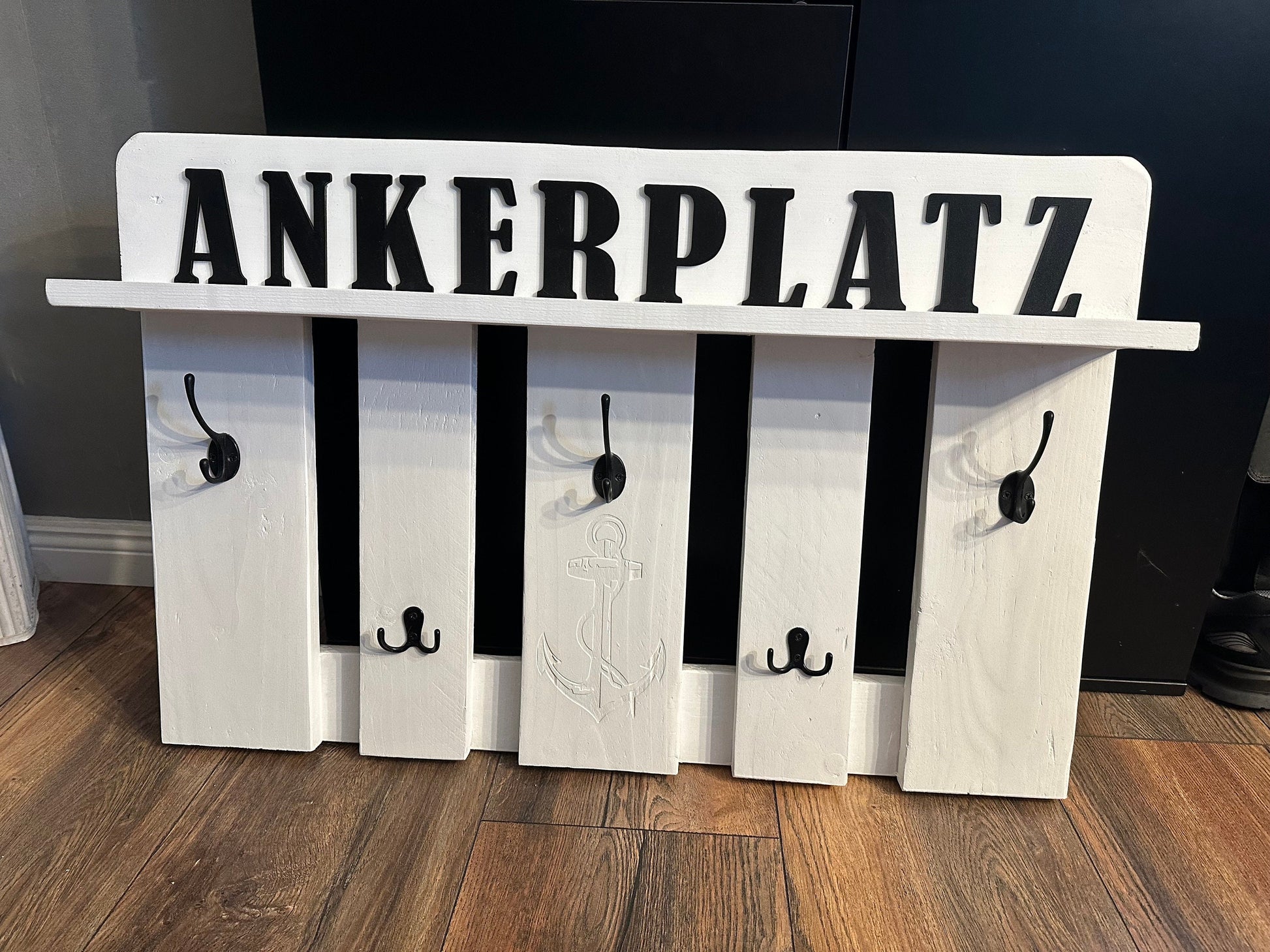Wall-mounted wooden coat rack with five vertical slats and multiple black
metal hooks. The top board features the word “ANKERPLATZ” (“anchor place” in
German) in bold black letters, with a central anchor engraving reinforcing the
nautical theme. The rack rests on a wooden floor, evoking rustic coastal
charm and practical entryway organization.