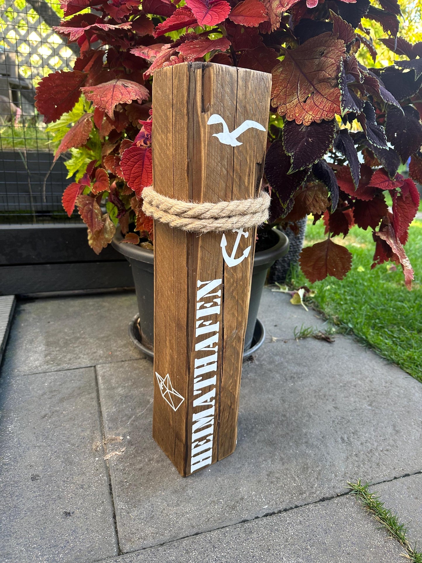Decorative wooden post placed on a paved patio beside a vibrant red and
green potted plant. The post features white nautical graphics including a
seagull, anchor, and paper boat, with thick rope wrapped around the top. The word “HEIMATHAFEN” (“home port” in German) is vertically printed in white,
evoking themes of coastal comfort, belonging, and maritime charm.